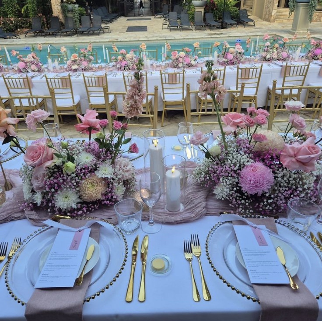 Table detail with blush florals, gold-rimmed plates and taper candles by a pool
